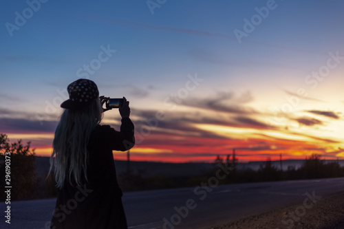 Back view of blonde girl, making photo of sunset with smartphone.