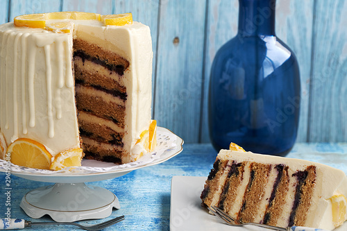 Front view of piece of layer cake with cream cheese, lemon and blueberries. In the background you can see the cake ans a blue bottle on a blue background.
