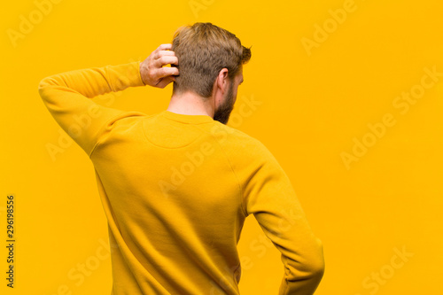 young blonde man thinking or doubting, scratching head, feeling puzzled and confused, back or rear view against orange wall