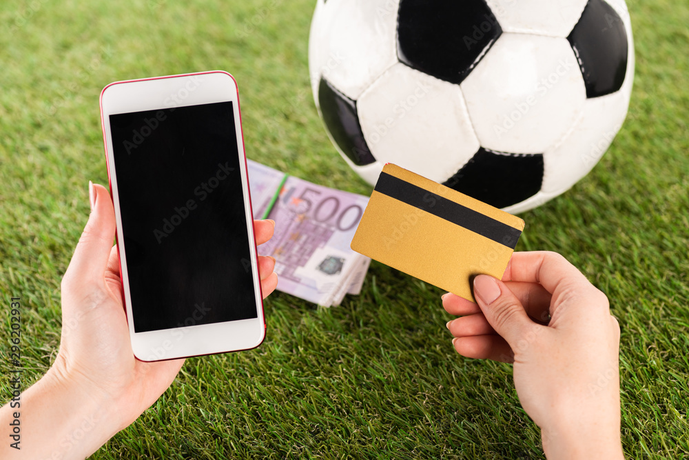 Fototapeta premium przycięty widok kobiecych rąk smartfonem i kartą kredytową w pobliżu piłki nożnej i banknotów euro na zielonej trawie, koncepcja zakładów sportowych