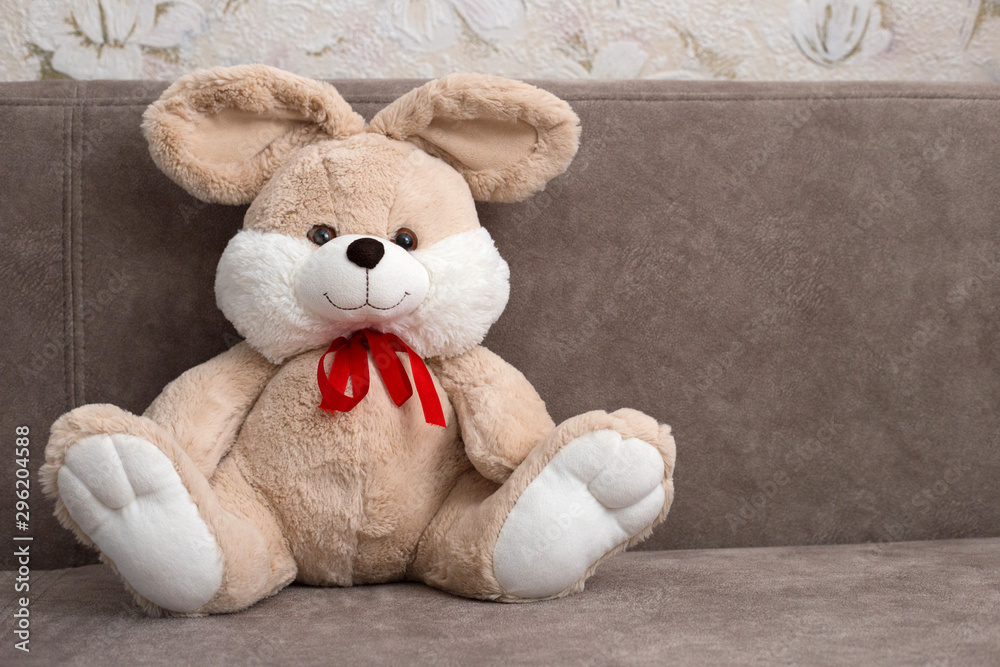 Fototapeta premium Plush rabbit sitting on a brown sofa. Beige toy rabbit with a red bow. Place for text. Romantic card