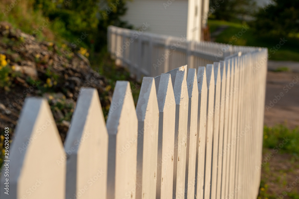 A white picket fence divides a garden from the road. A white building can be seen in the background. The sun is shinning on the fence illuminating the textured wood.