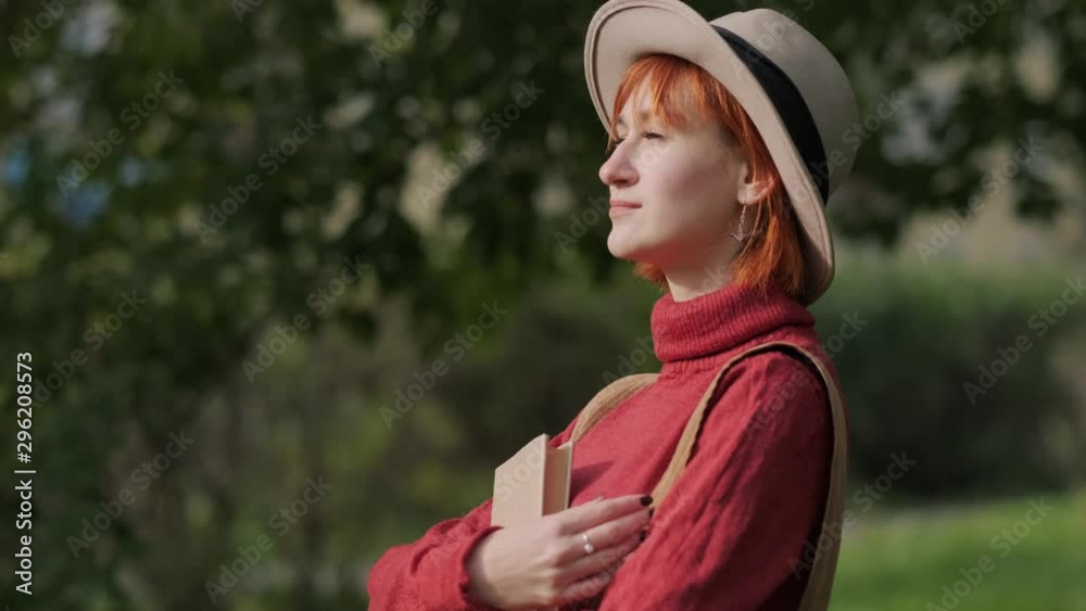 Young attractive redhead woman in a hat and sweater reading a book on a bench in a natural park. Autumn cool day, green and yellow trees. Young attractive redhead woman in a hat and sweater reading a book on a bench in a natural park. Autumn cool day, green and yellow trees.