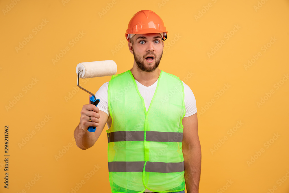 Handy man with paint roller, paints walls at home, does renovation and repairing, wears helmet, apron, busy with room painting job, holds special tool, looks with embarrassement. Building concept
