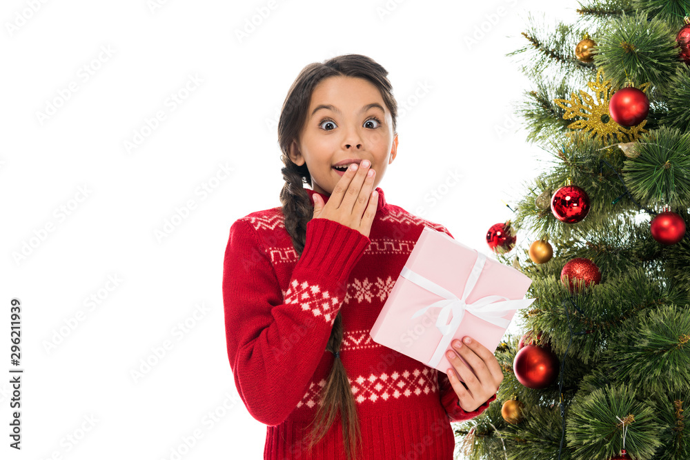 excited child covering face and holding pink present near christmas tree isolated on white