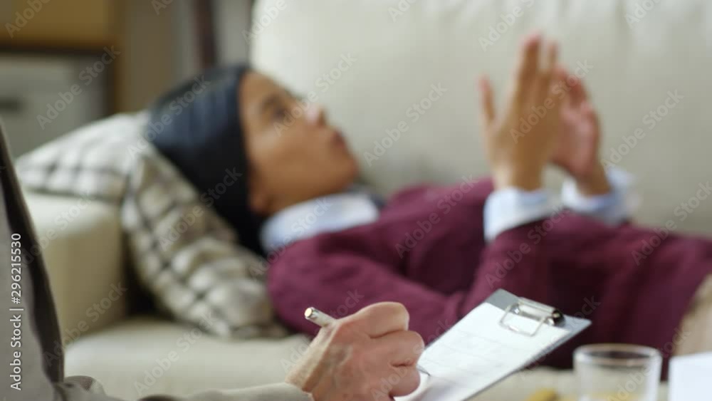 Close up shot of unrecognizable female psychologist taking notes on clipboard while listening to woman lying on sofa during therapy session; focus in the foreground Close up shot of unrecognizable female psychologist taking notes on clipboard while listening to woman lying on sofa during therapy session; focus in the foreground