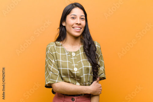 young pretty hispanic woman laughing shyly and cheerfully, with a friendly and positive but insecure attitude against brown wall photo
