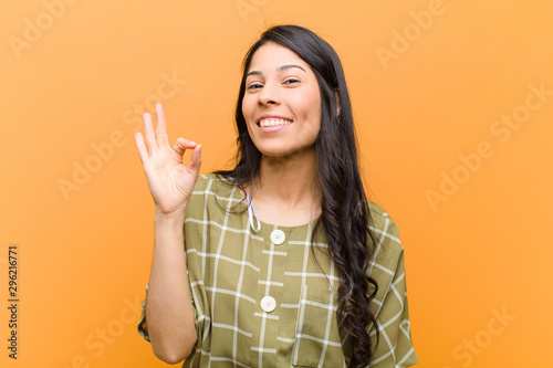 young pretty hispanic woman feeling happy, relaxed and satisfied, showing approval with okay gesture, smiling against brown wall photo