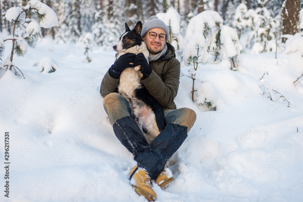Fototapeta premium Happy man holding lovely dog in his hands in snowy forest. Smiling boy hugging adorable puppy in winter wood. Pet lover. Dog - human`s friend concept.