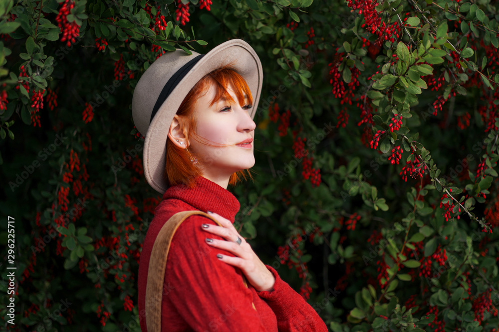 Portrait of a young attractive girl with red hair in a hat and a red sweater against the background of a bush of barberry with red berries. Portrait of a young attractive girl with red hair in a hat and a red sweater against the background of a bush of barberry with red berries.