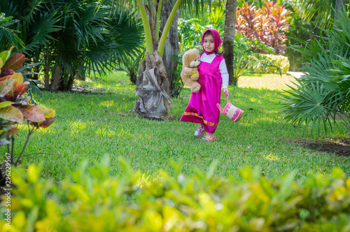 Pretty little girl in pink dress with headscarf plays and walks through the garden  flat green grass and trees