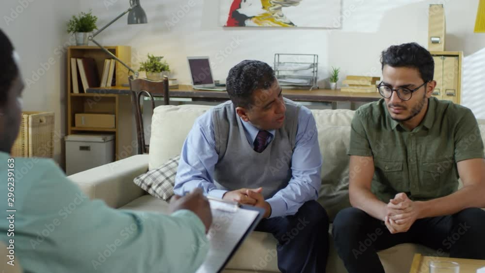 Middle aged arab man sitting with adult son on sofa and talking with male african american psychotherapist during counseling session