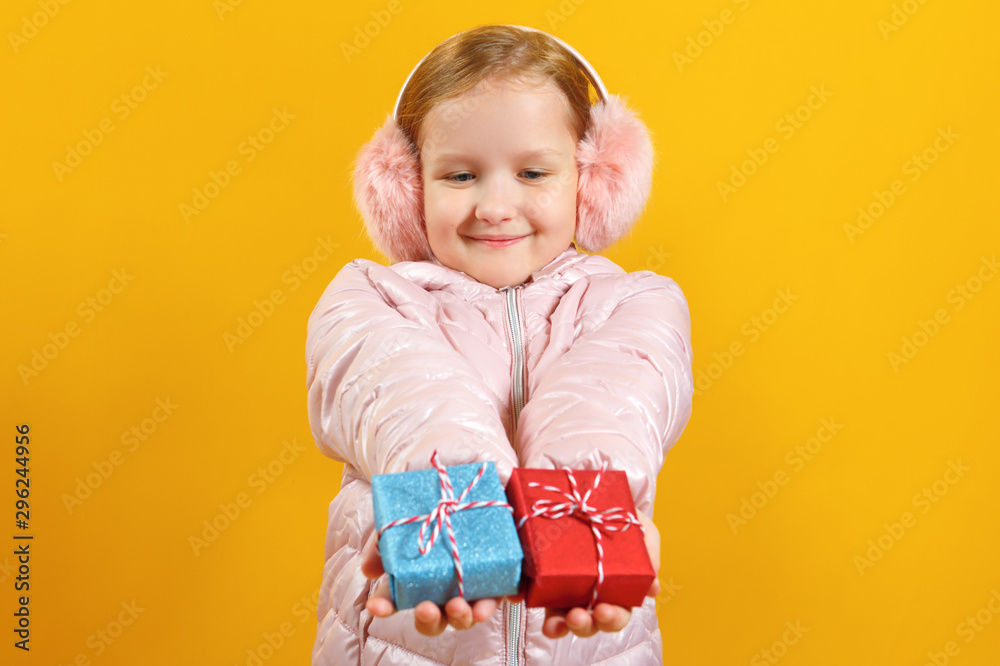 Happy little girl holds out small boxes with gifts. A child in a jacket and warm ear muffs on a yellow background. Christmas and new year concept Happy little girl holds out small boxes with gifts. A child in a jacket and warm ear muffs on a yellow background. Christmas and new year concept