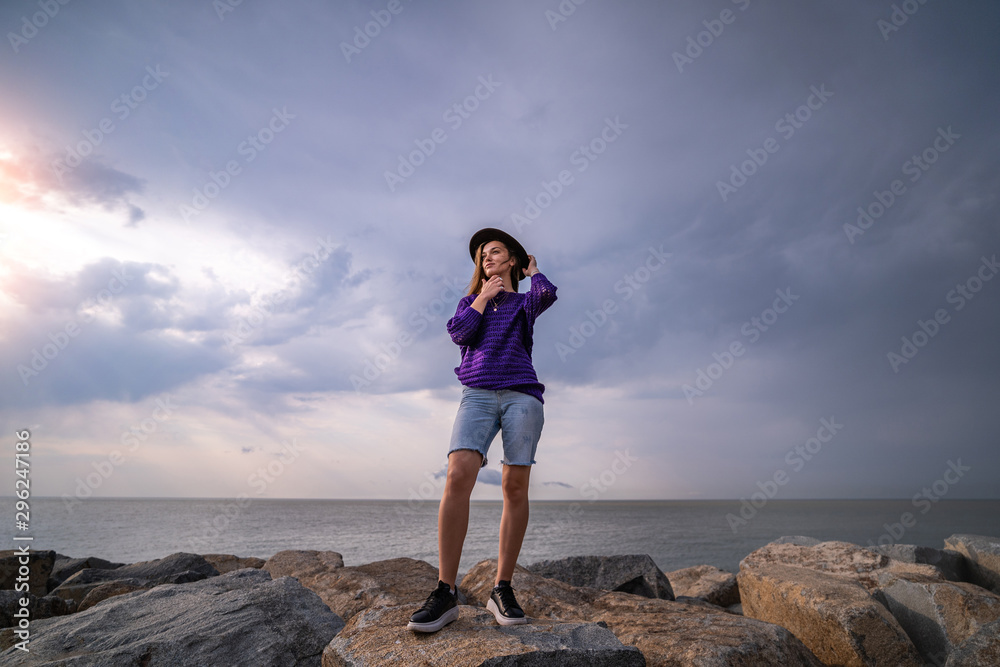 Attractive young thoughtful woman traveler in a hat stands on a rocky shore on a background of moody sky and black sea