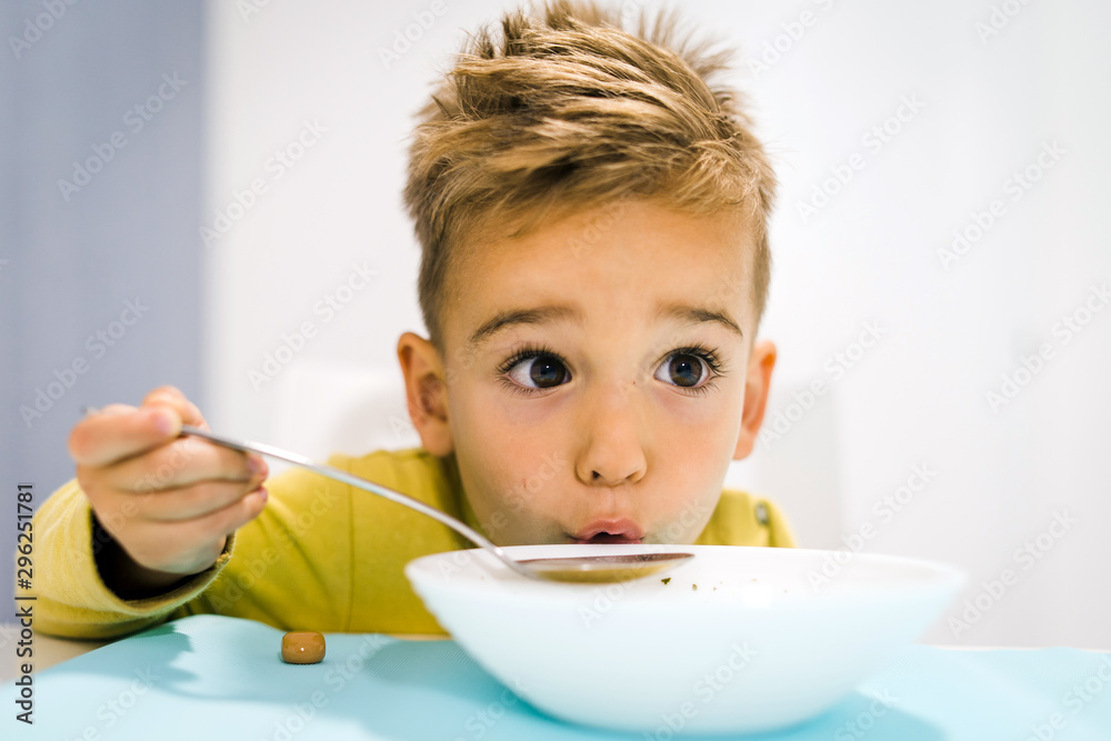 Portrait of small boy child eating soup meal or breakfast having lunch by the table at home with spoon white kindergarten Portrait of small boy child eating soup meal or breakfast having lunch by the table at home with spoon white kindergarten
