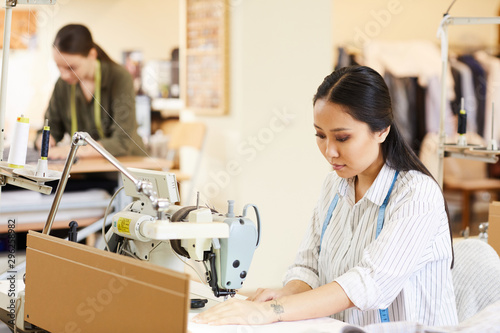 Asian dressmaker sitting at the table and using sewing machine for sewing clothes in atelier