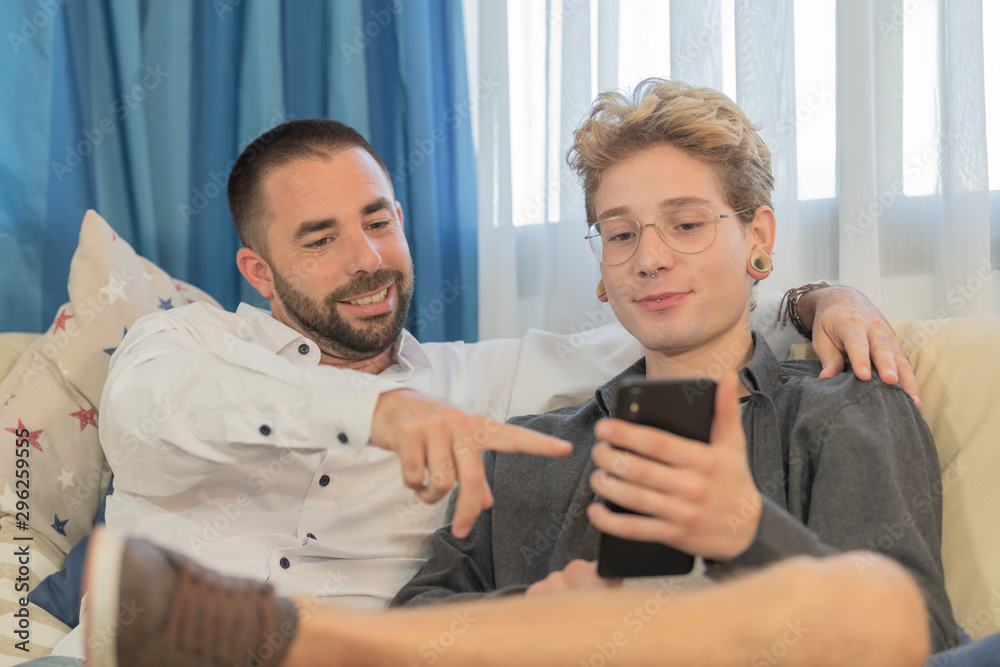 A bearded man points to a photo on his friend's smartphone with blond hair and glasses, both sitting on the sofa, relaxing. Concept of coexistence and sociability. A bearded man points to a photo on his friend's smartphone with blond hair and glasses, both sitting on the sofa, relaxing. Concept of coexistence and sociability.