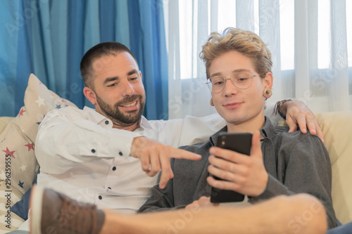 A bearded man points to a photo on his friend's smartphone with blond hair and glasses, both sitting on the sofa, relaxing. Concept of coexistence and sociability.