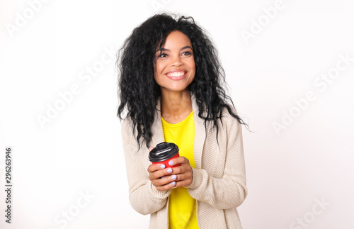 Coffee break. Happy African ethnic woman in beige cardigan and yellow t-shirt is holding a red paper coffee cup and smiling while looking to the right.