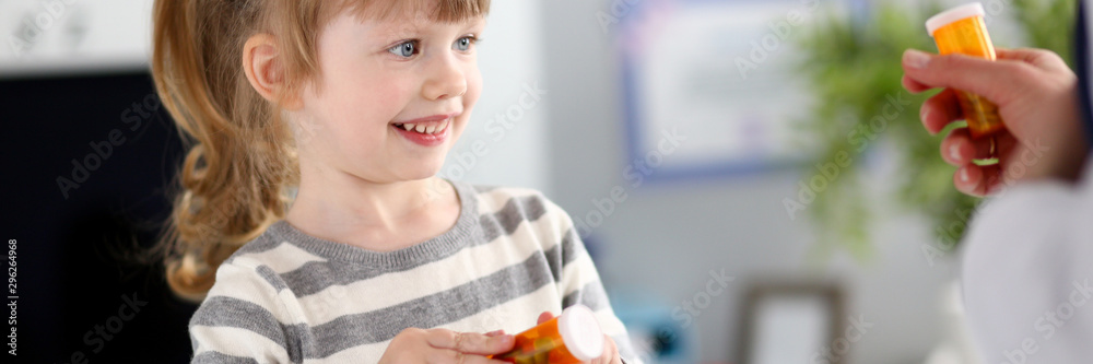 Portrait of small girl sitting in big modern doctor office and talking with pediatrician. Joyous child in striped outfit holding container with pills. Medicine and healthcare concept Portrait of small girl sitting in big modern doctor office and talking with pediatrician. Joyous child in striped outfit holding container with pills. Medicine and healthcare concept