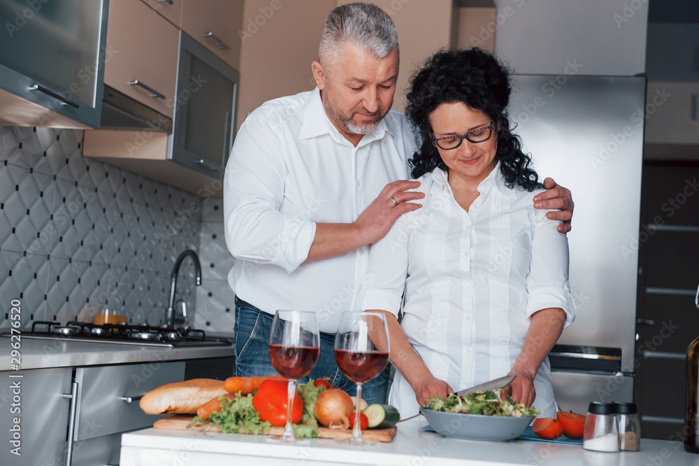 Happy together. Man and his wife in white shirt preparing food on the kitchen using vegetables Happy together. Man and his wife in white shirt preparing food on the kitchen using vegetables