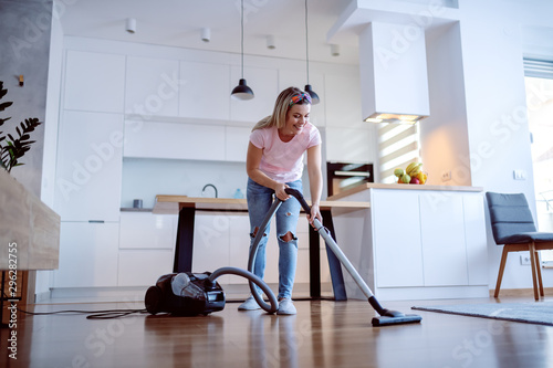 Smiling cheerful caucasian blonde housewife using vacuum cleaner to clean floor in living room. Home interior.