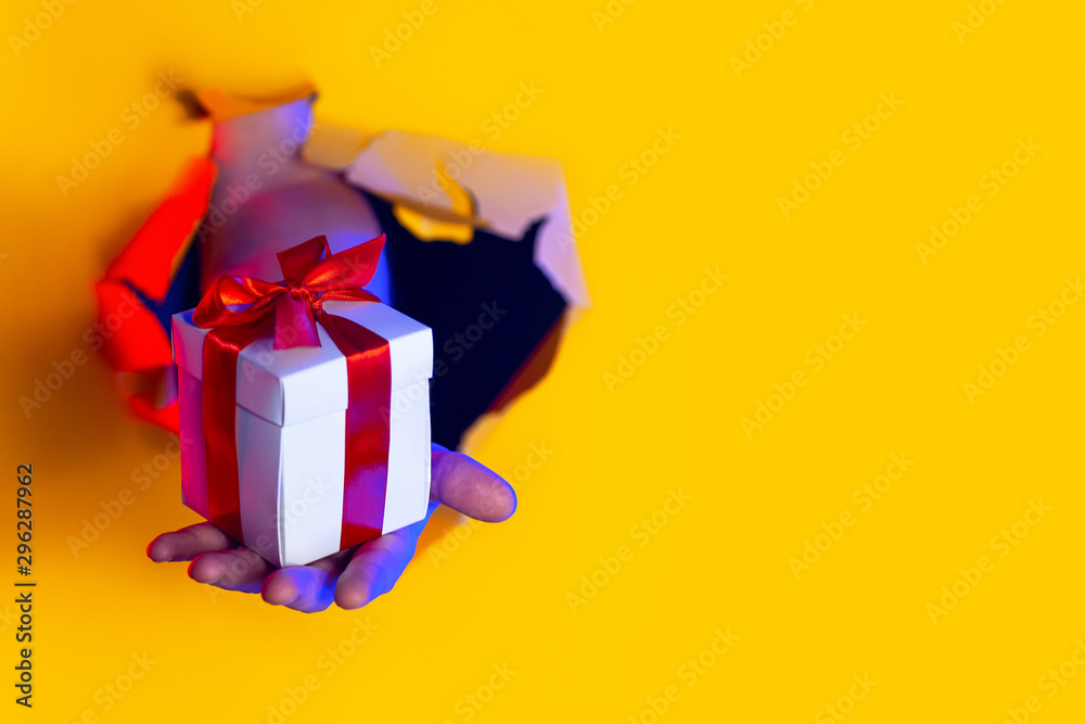 A gift with a red bow in hand emerges from a ragged hole in yellow paper background, illuminated by neon light