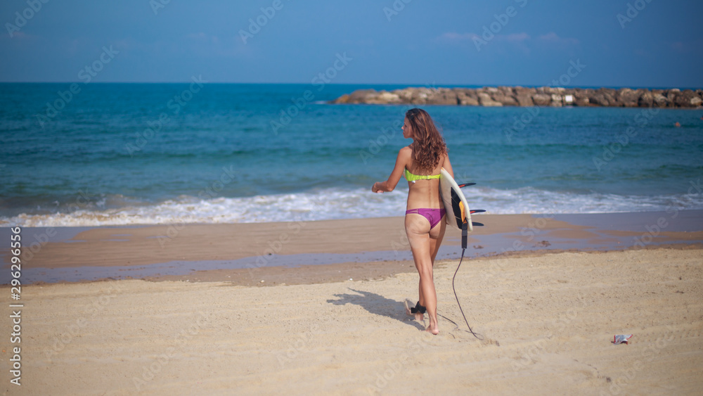Athletic woman surfer, holding her surfboard under her arm and walking towards the sea. Breakewater in the far background. Athletic woman surfer, holding her surfboard under her arm and walking towards the sea. Breakewater in the far background.
