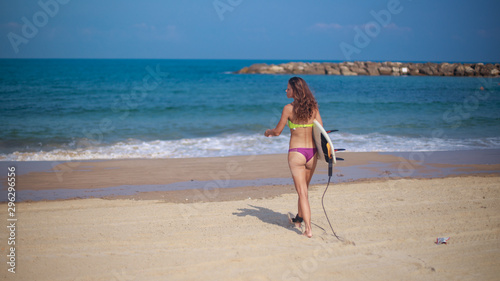 Athletic woman surfer, holding her surfboard under her arm and walking towards the sea. Breakewater in the far background.