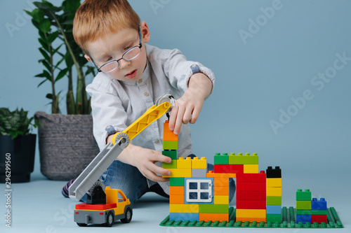 Little ginger boy sitting on floor assembling house from construction toy blocks