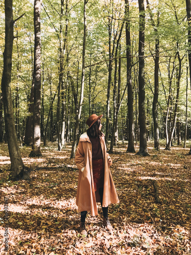 Hipster woman in hat and coat traveling in autumn sunny forest with fall leaves. Stylish traveler girl exploring autumn woods, enjoying sunny day © sonyachny