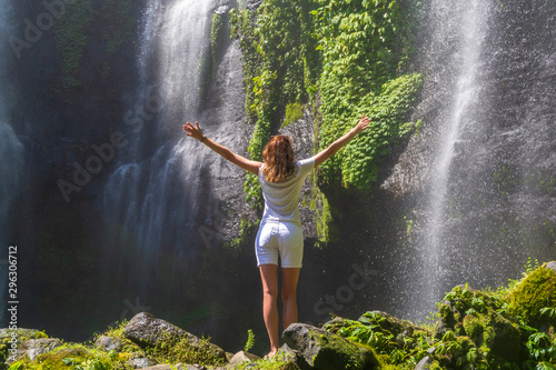 A young woman stands with her back in white clothes at the Sekumpul Great Waterfall in the deep rainforest of Bali Island, Indonesia.