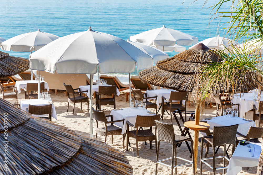 Restaurant tables on a sandy beach under thatched roof umbrellas. Relax and service at a fashionable resort.