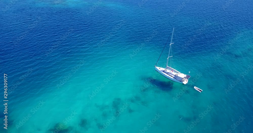 Panning aerial view of a white sailboat with an inflatable boat attached to the back floating in clear turquoise blue water in the Caribbean Panning aerial view of a white sailboat with an inflatable boat attached to the back floating in clear turquoise blue water in the Caribbean