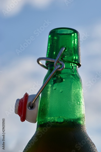 A selective-focus macro image of condensation - glistening enticingly in the afternoon sunshine - on a green-glass swing-top beer bottle; with a lightly clouded blue sky, blurry background. photo