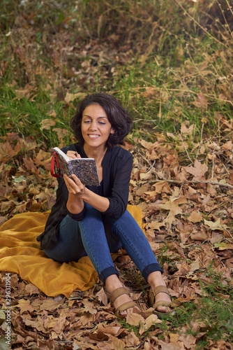  Latin brunette woman with a black t-shirt in a forest with dry leaves and autumnal colors, the sun enters between the branches of the trees, she is sitting with a notebook in her hands.