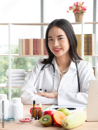 Young Asian female nutritionist testing food samples and writing records on a clipboard. Fresh fruits, vitamins and medicines on table in laboratory. Nutrition, healthy eating and diet concept.