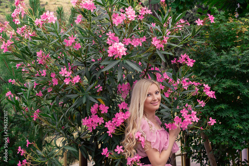 Beautiful young blonde woman in a pink dress stands posing outdoors near a bush with flowers.