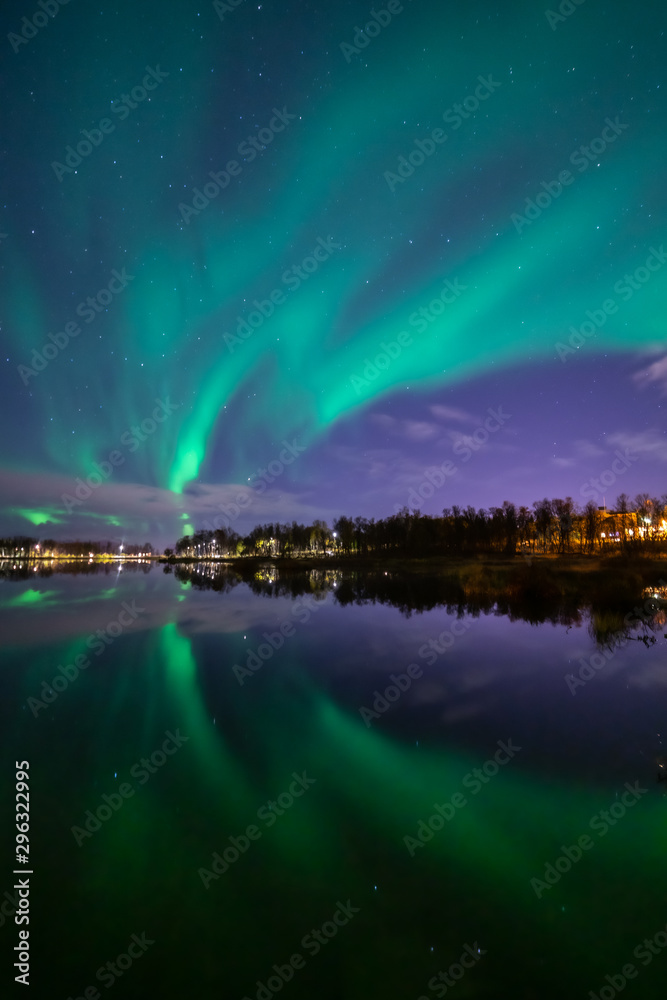 Northern lights above lake. Green aurora on purple sky with stars and clouds. Trees, city light. Reflections in water. Prestvannet, Tromso, Norway.
