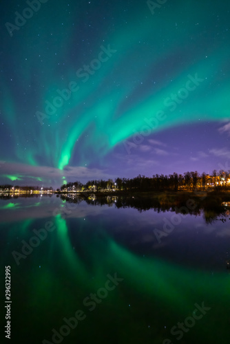 Northern lights above lake. Green aurora on purple sky with stars and clouds. Trees  city light. Reflections in water. Prestvannet  Tromso  Norway.