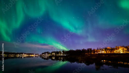Northern lights above lake. Green aurora on purple sky with stars and clouds. Trees, city light. Reflections in water. Prestvannet, Tromso, Norway.