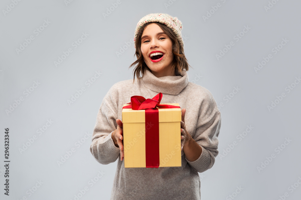 Fototapeta premium christmas, holidays and people concept - happy smiling young woman in knitted winter hat and sweater holding gift box over grey background