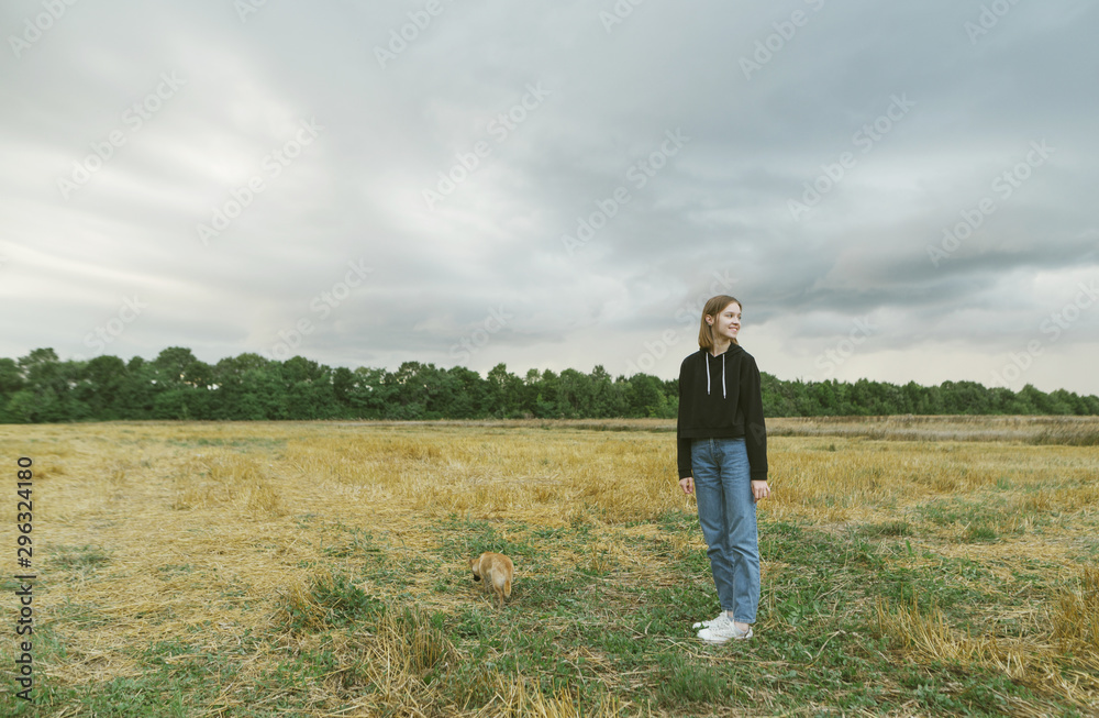 Cute girl in casual clothes walking with a small dog in the autumn field, looking away and smiling. Field portrait of a full-grown girl with a dog.