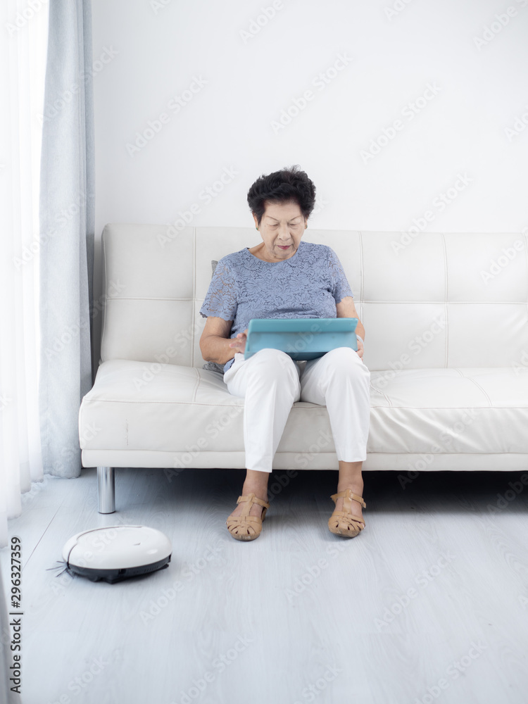 Senior woman unsing tablet while robot vacuum cleaning floor at home. Modern lifestyle concept.