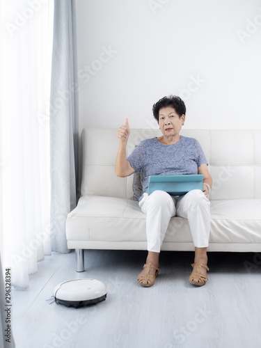 Senior woman unsing tablet while robot vacuum cleaning floor at home. Modern lifestyle concept.