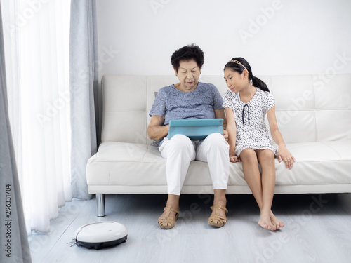 Asian senior woman and girl unsing tablet while robot vacuum cleaning floor at home. Modern lifestyle concept.