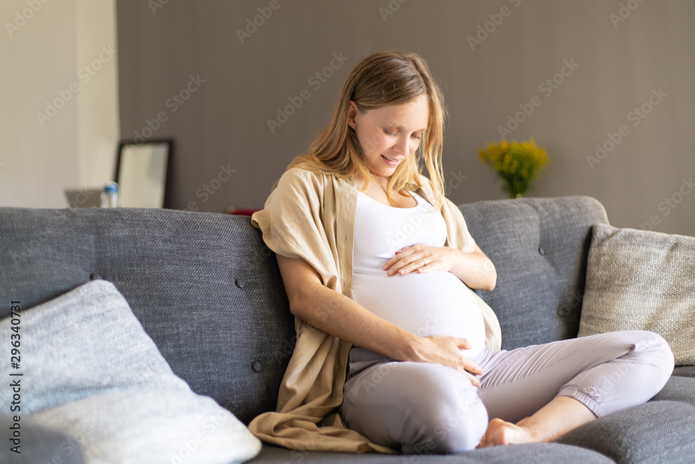 Excited expectant mother feeling baby moving inside stomach. Young pregnant woman sitting on couch, touching belly. Enjoying pregnancy concept