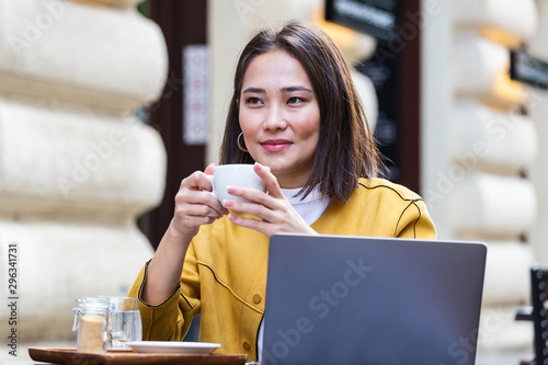 Young asian business woman sitting at table in cafe shop and writing in notebook. On table is laptop  smartphone and she is holding cup of coffee. Freelancer working in coffee shop.