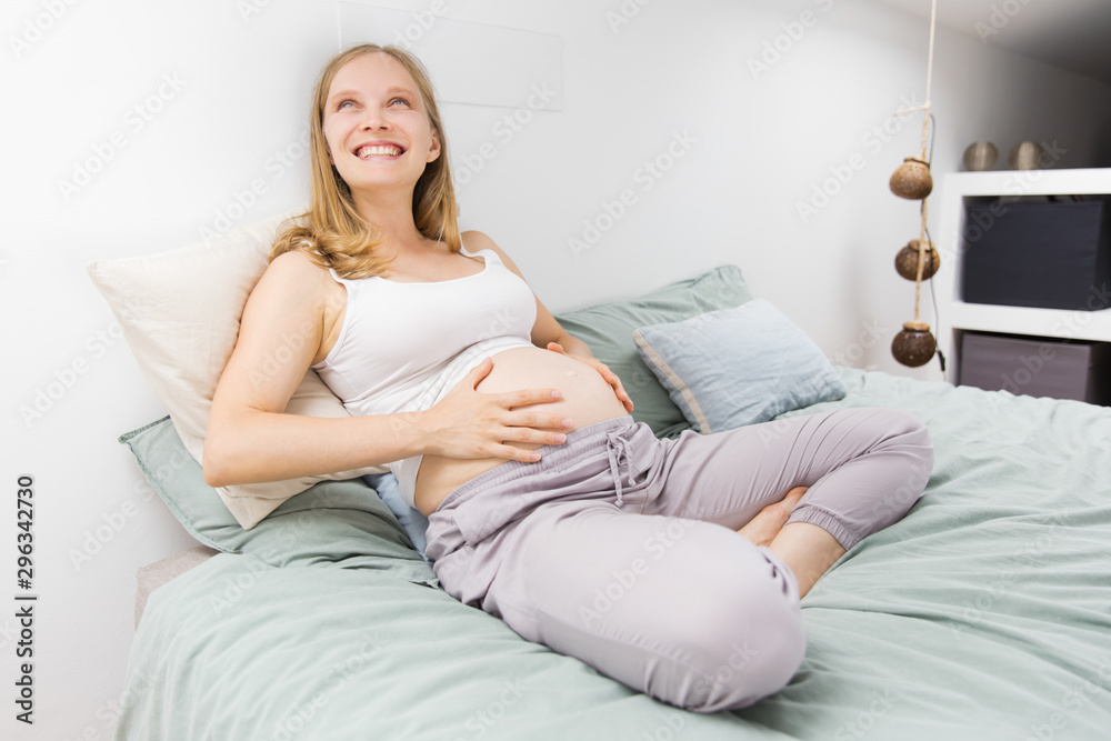 Joyful expectant mother resting in bed at home. Young pregnant woman holding belly, looking up, smiling, laughing. Happy future mother concept Joyful expectant mother resting in bed at home. Young pregnant woman holding belly, looking up, smiling, laughing. Happy future mother concept