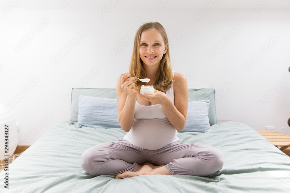 Cheerful expectant mother keeping healthy diet. Pregnant woman sitting on bed and eating yogurt. Milk food and pregnancy concept Cheerful expectant mother keeping healthy diet. Pregnant woman sitting on bed and eating yogurt. Milk food and pregnancy concept